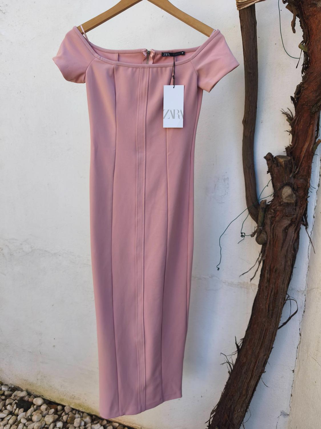 Pale pink, off-the-shoulder dress hanging on a wooden hanger, with a visible "ZARA" tag. White wall background.