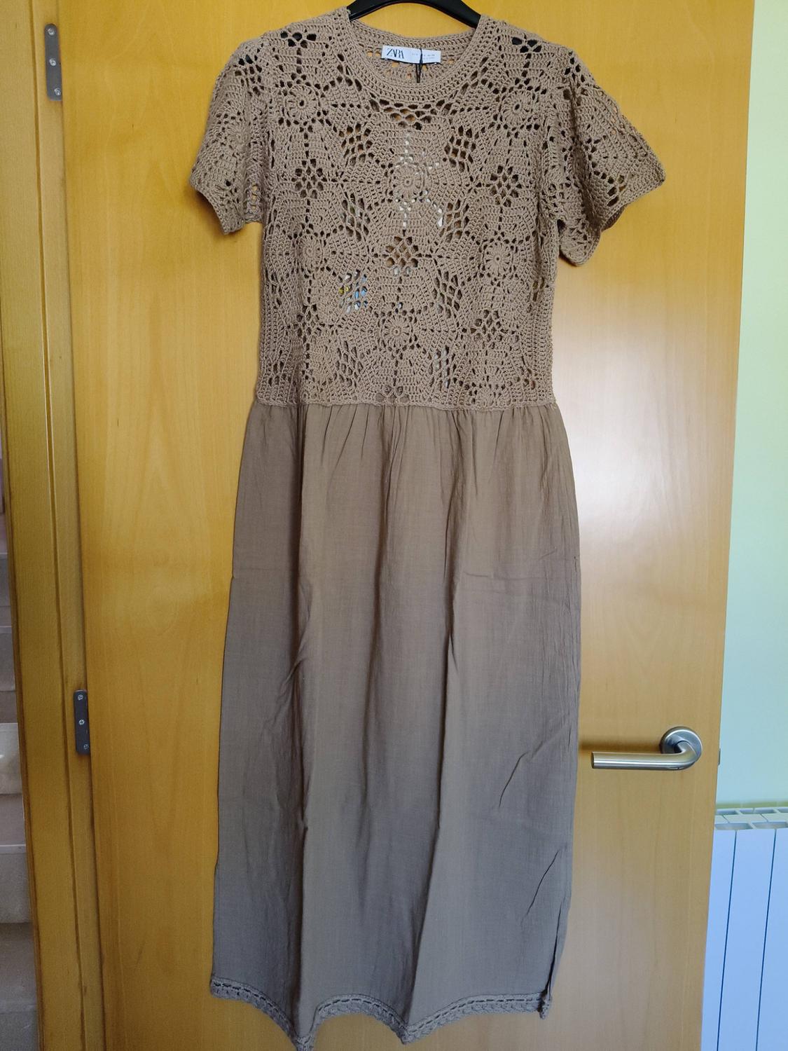 Beige dress hanging on a wooden door. Openwork crochet bodice, short sleeves and plain skirt. ZARA label. Casual setting.