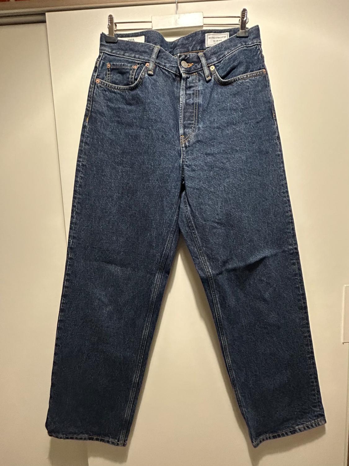 Dark blue jeans hanging on a hanger against a white background. Classic design with five pockets and a button closure.