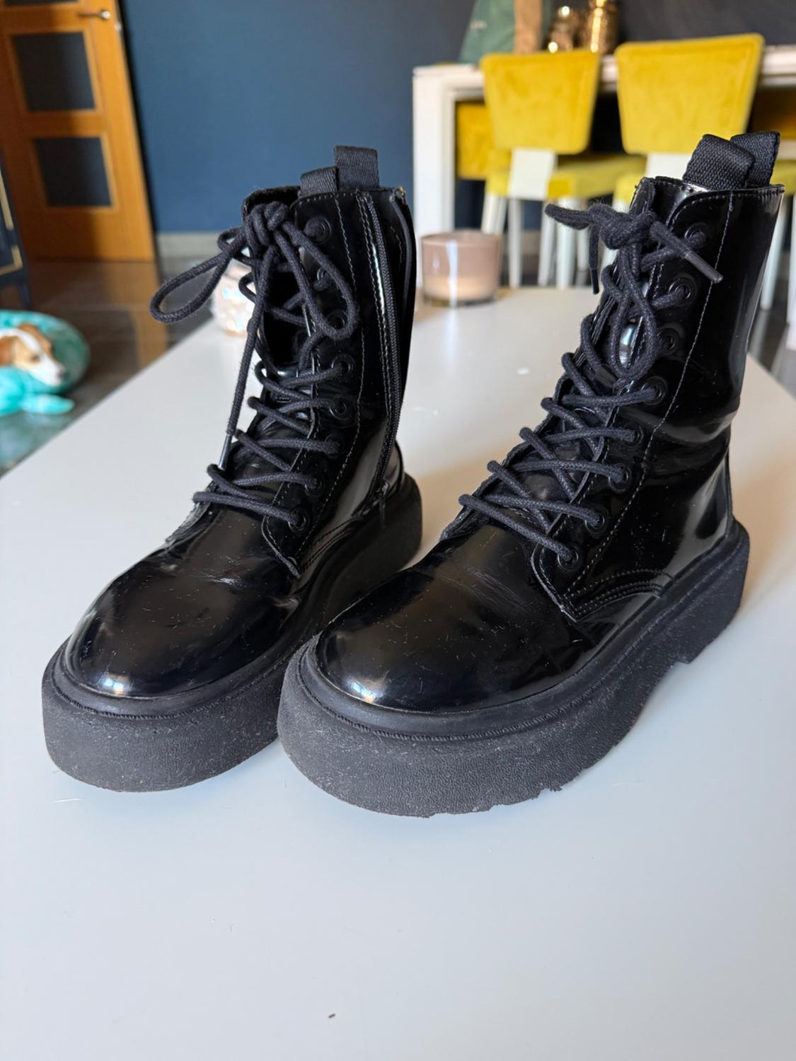 Black patent leather platform boots with laces, on a white table. Background with yellow chairs and blue wall.