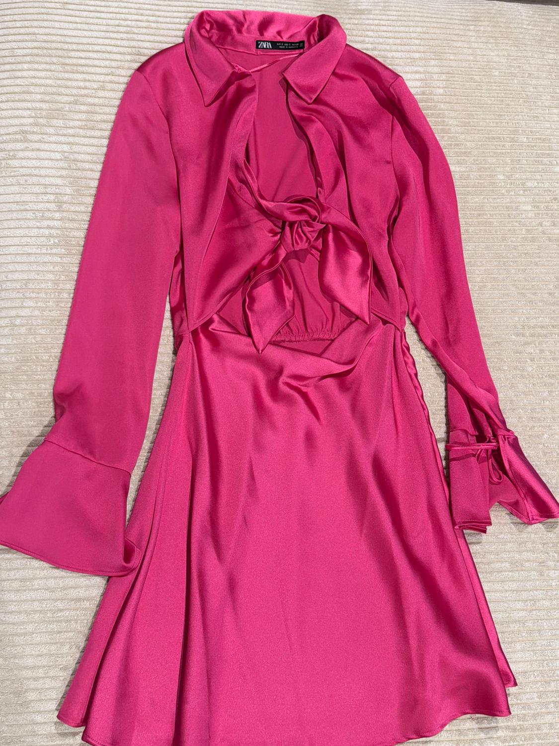 Satin fuchsia pink short dress, long flared sleeves, neckline and front knot with an opening, against a light textured background. Text: ZARA.