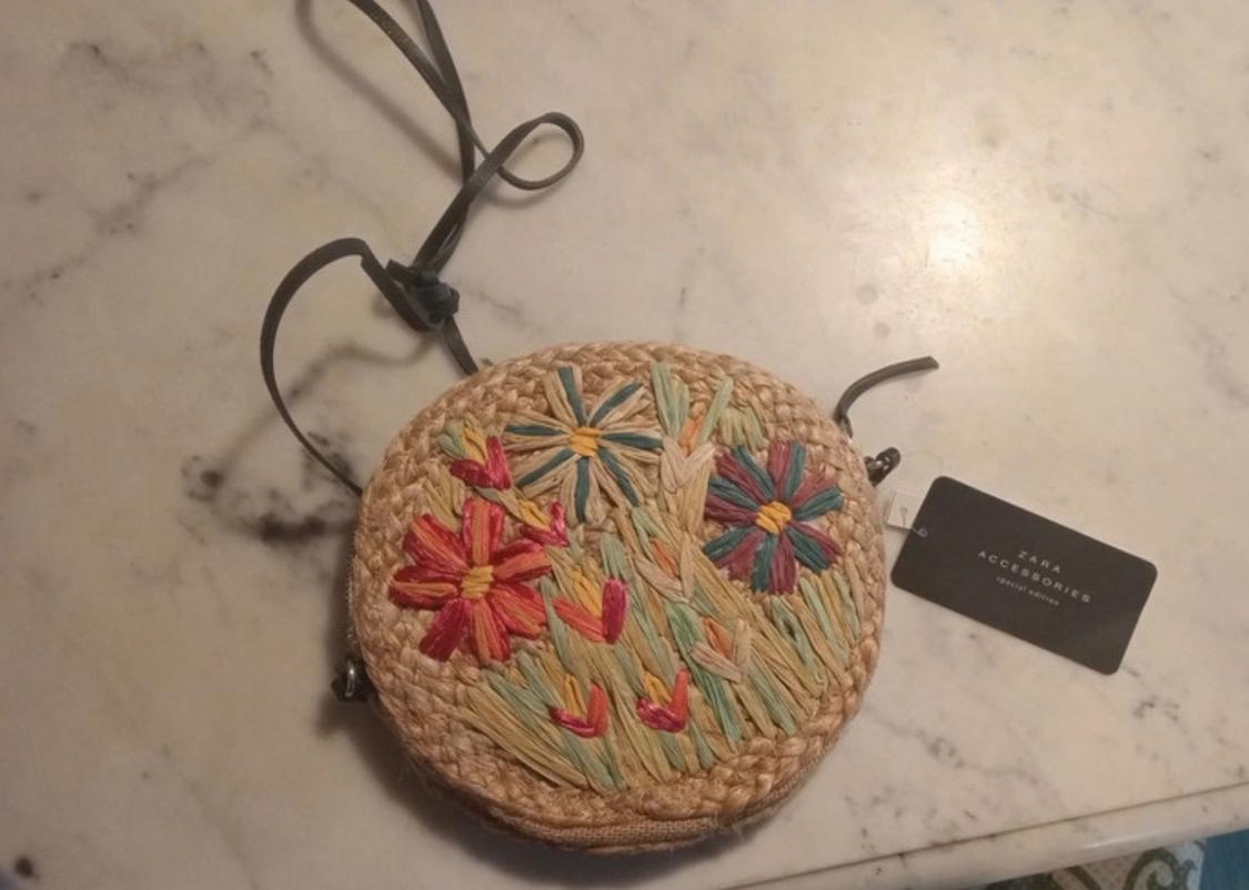 Round straw bag with brightly embroidered flowers on a marble table. Label: "ZARA ACCESSORIES".