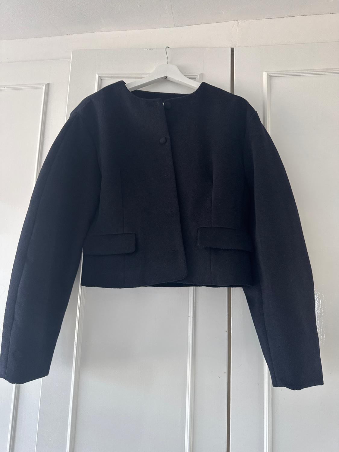 Black long-sleeved jacket hanging on a white hanger over a white door. Minimalist and elegant design.