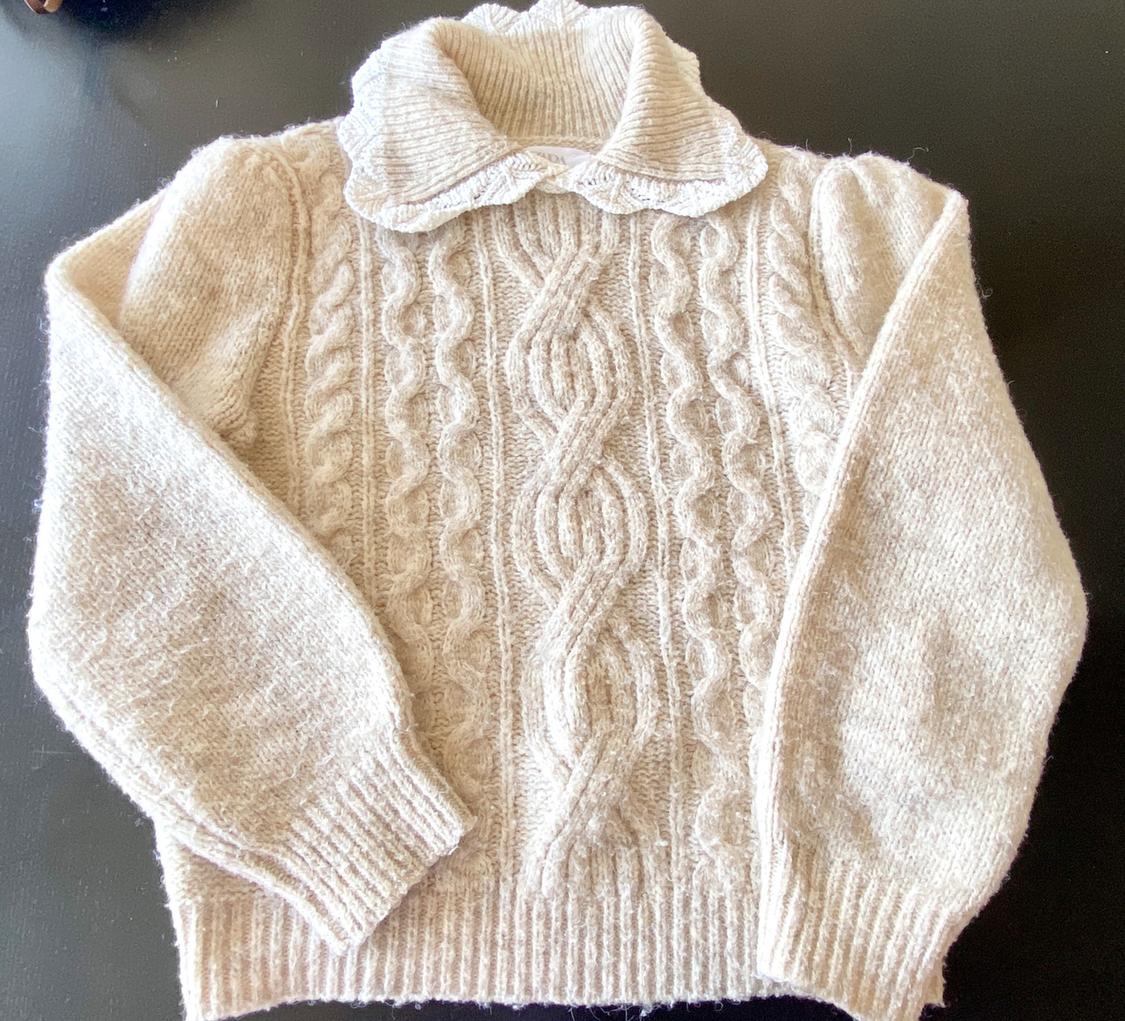 Beige knitted jumper with cable knit design and roll neck on a black background. Long sleeves and visible soft texture.