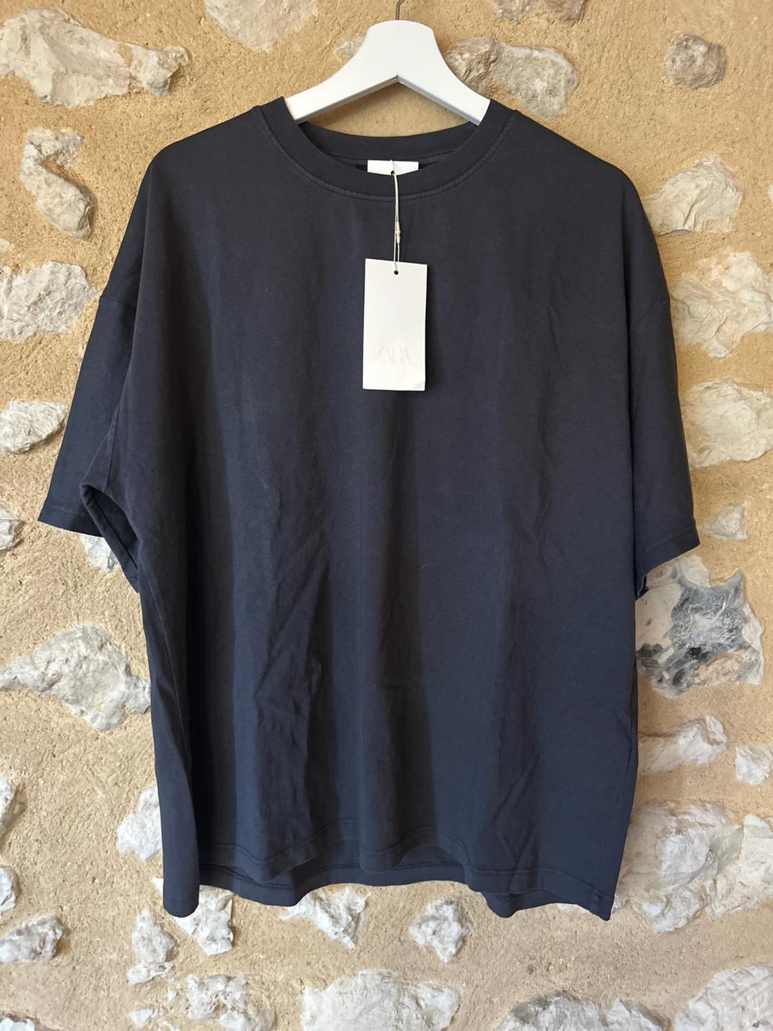 Dark grey short-sleeved T-shirt hanging on a white hanger against a stone wall background. Label with the text ZARA.