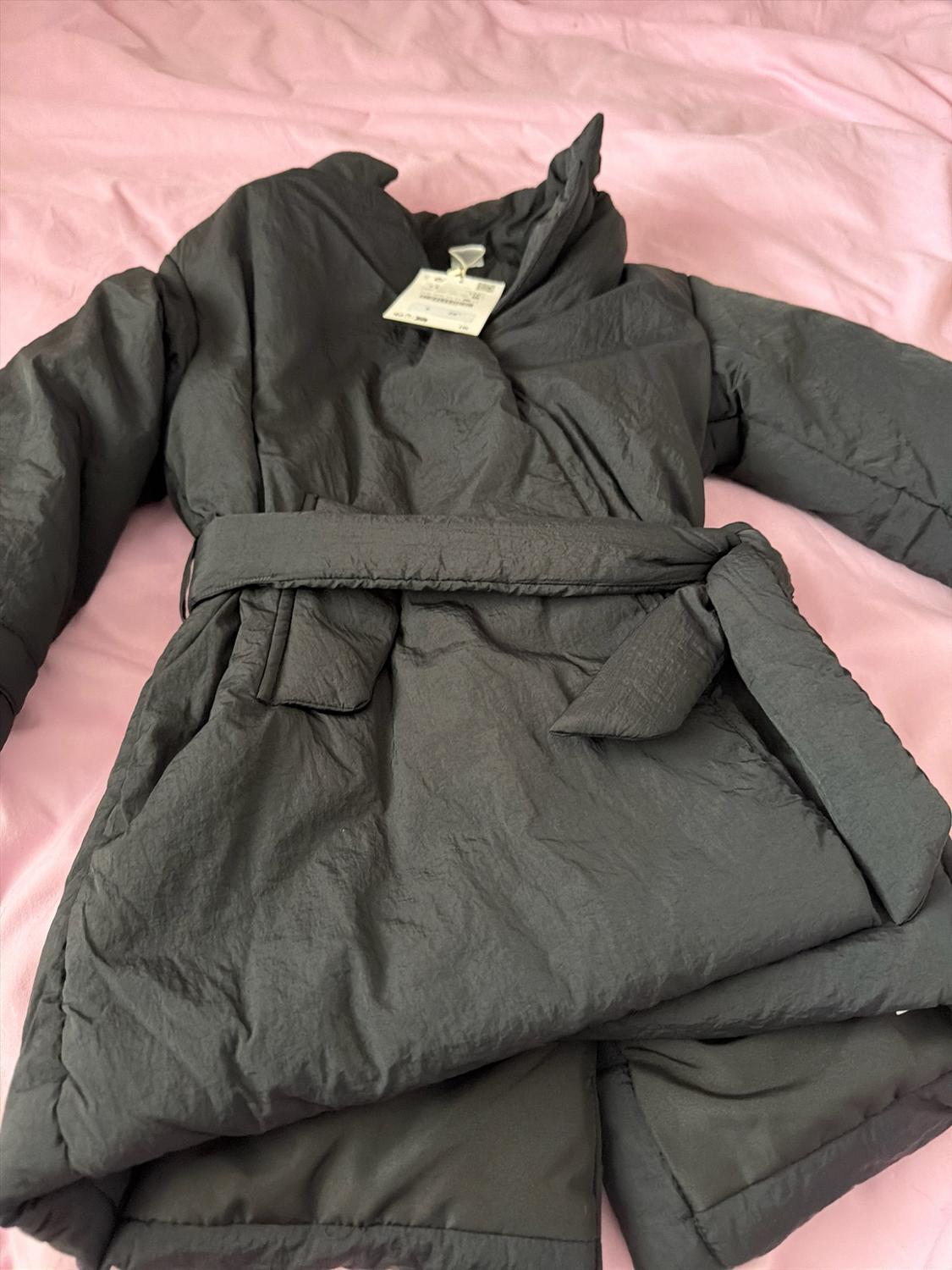 Black quilted coat with a tied belt, set against a crumpled pink fabric background. Label hanging from the collar.