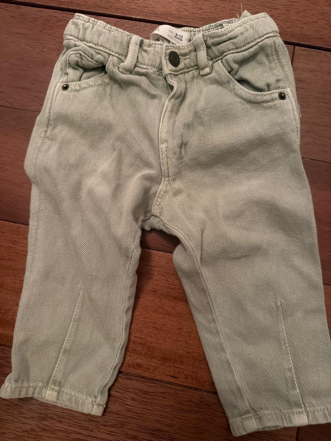 Light green baby pants on a wooden floor. They have front pockets and belt loops. Visible label on the waistband.