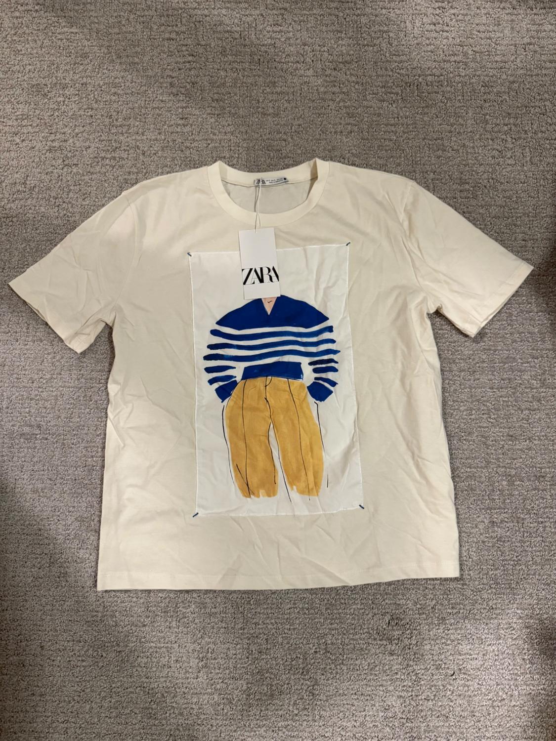 White t-shirt with a figure print in a blue sweater and brown pants. Hang tag with "ZARA" text. Gray carpet background.