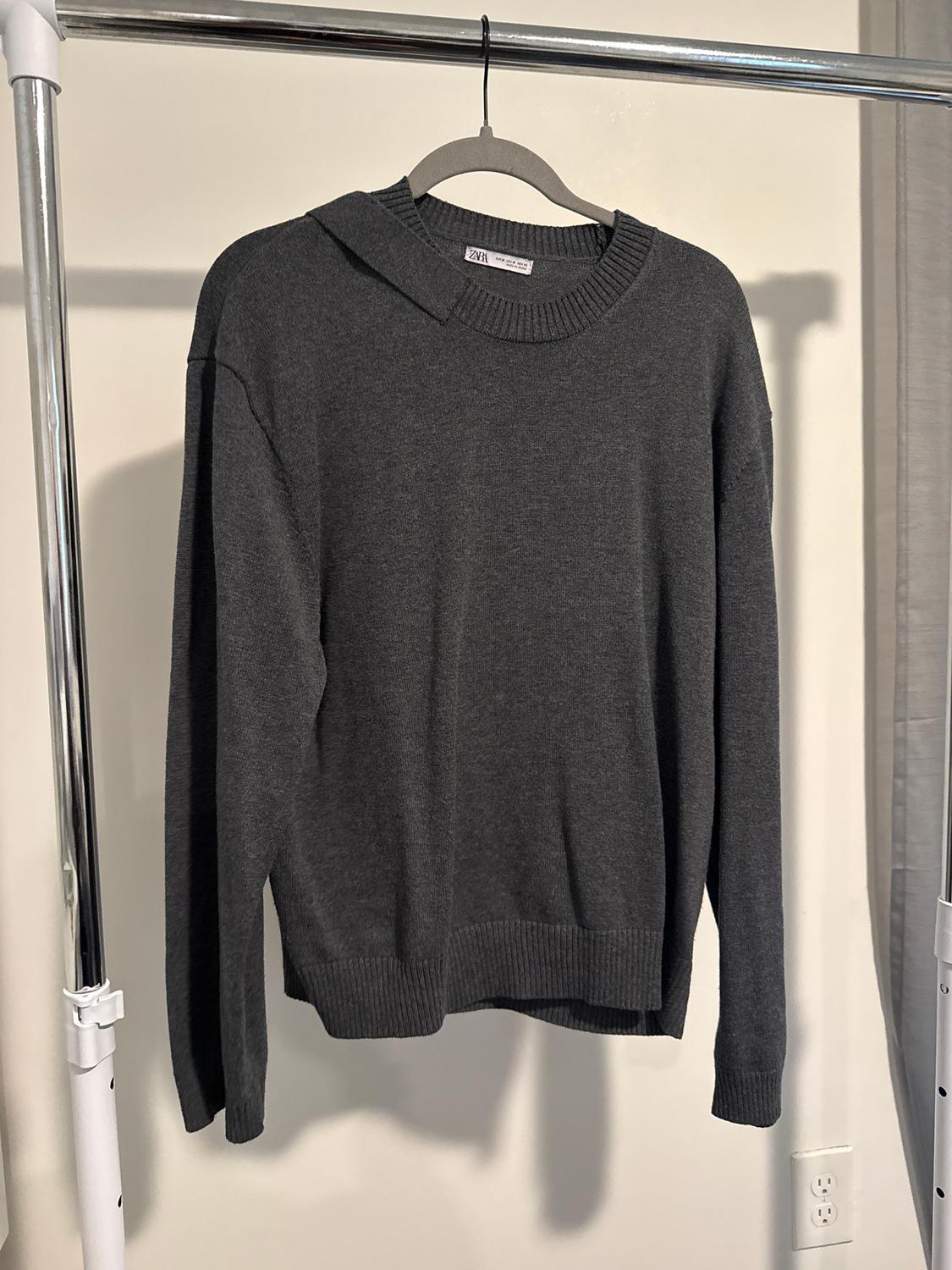 Dark grey jumper hanging on a metal hanger in a lit indoor setting. Visible label with text "ZARA".