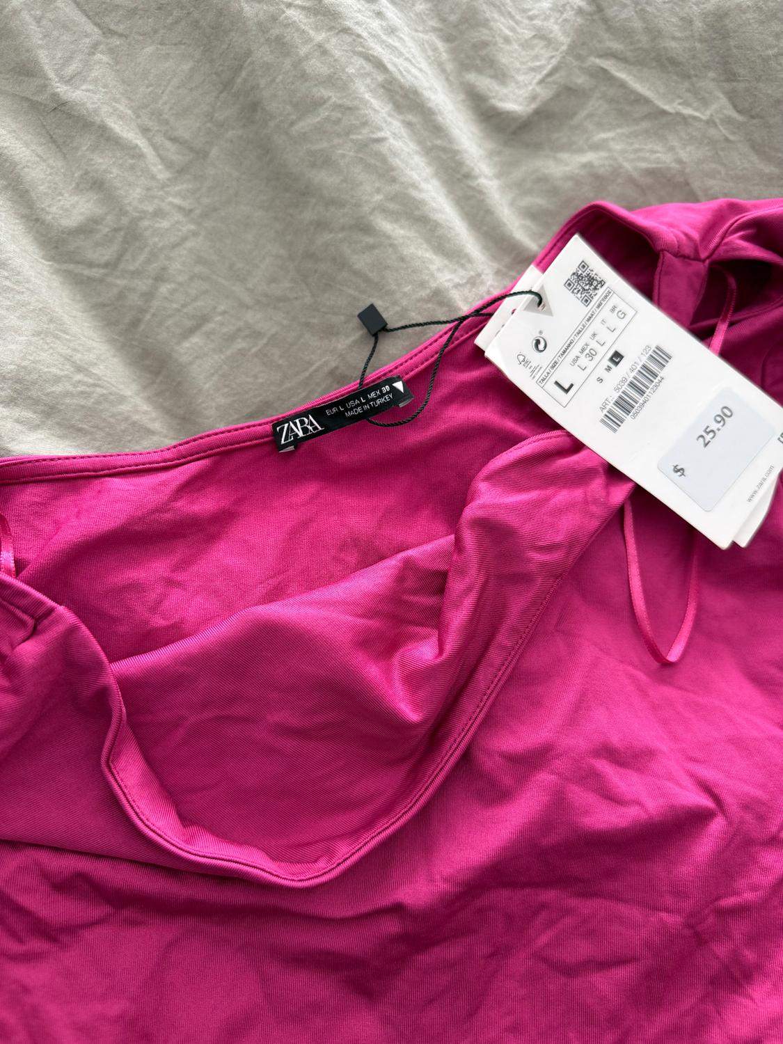 Zara pink garment tag, size L, made in Turkey, priced at $25.90, on a wrinkled beige fabric background.