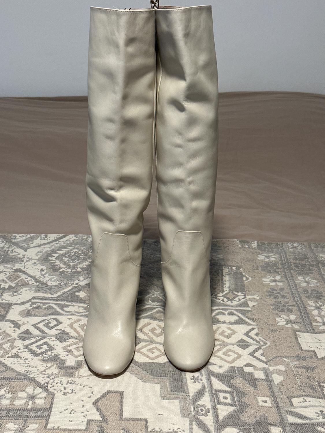 Beige knee-high boots on a rug with a geometric design in grey and beige tones. White wall background and light brown floor.