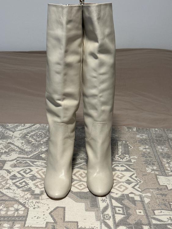 Beige high boots on a rug with a geometric design in gray and beige tones. White wall background and light brown floor.