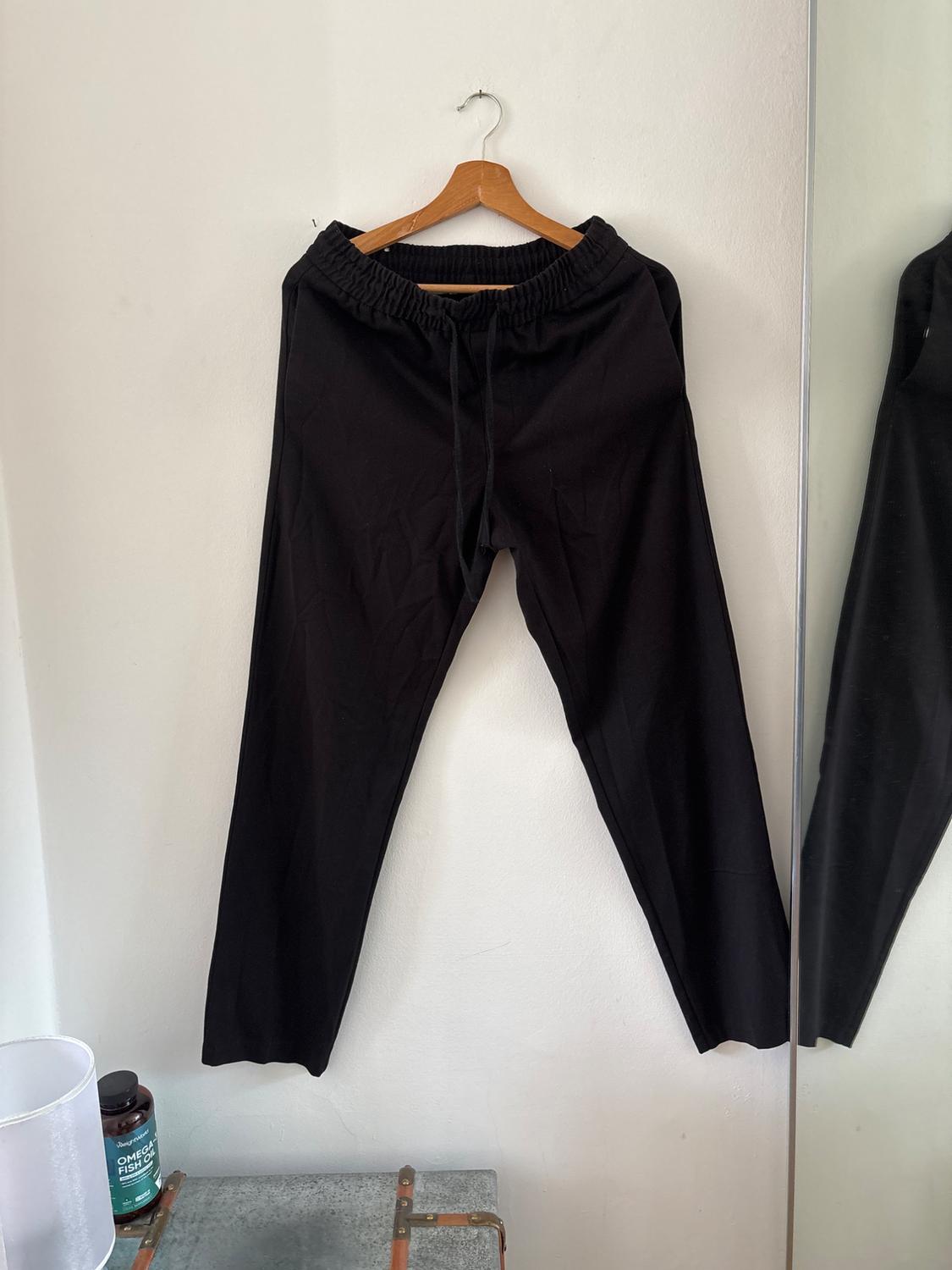 Black trousers with a drawstring hanging on a wooden hanger against a white wall next to a mirror. Below, a tub with the text OMEGA FISH OIL.