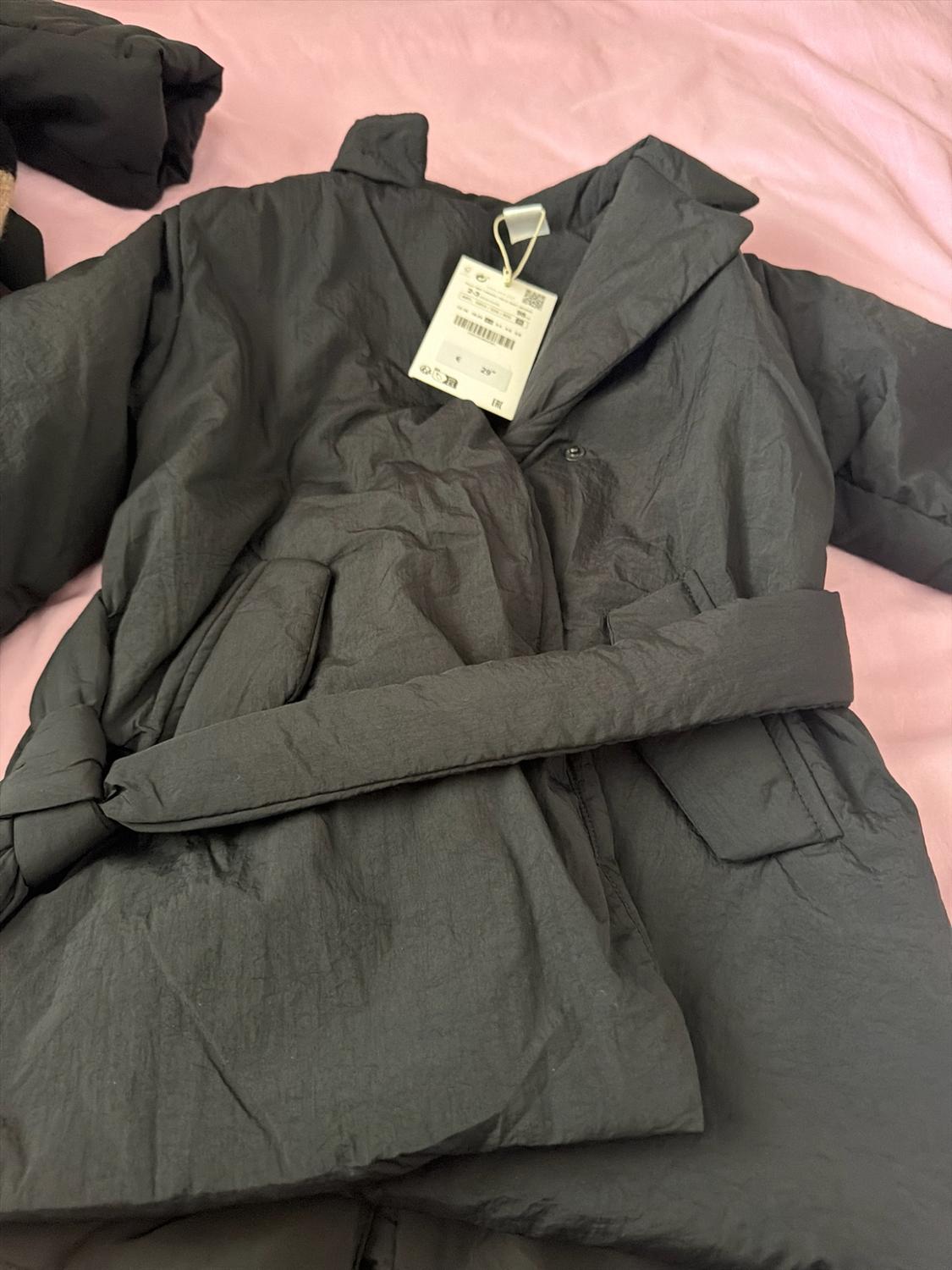 Black padded coat with a belt and visible label, laid out on a pink fabric background.
