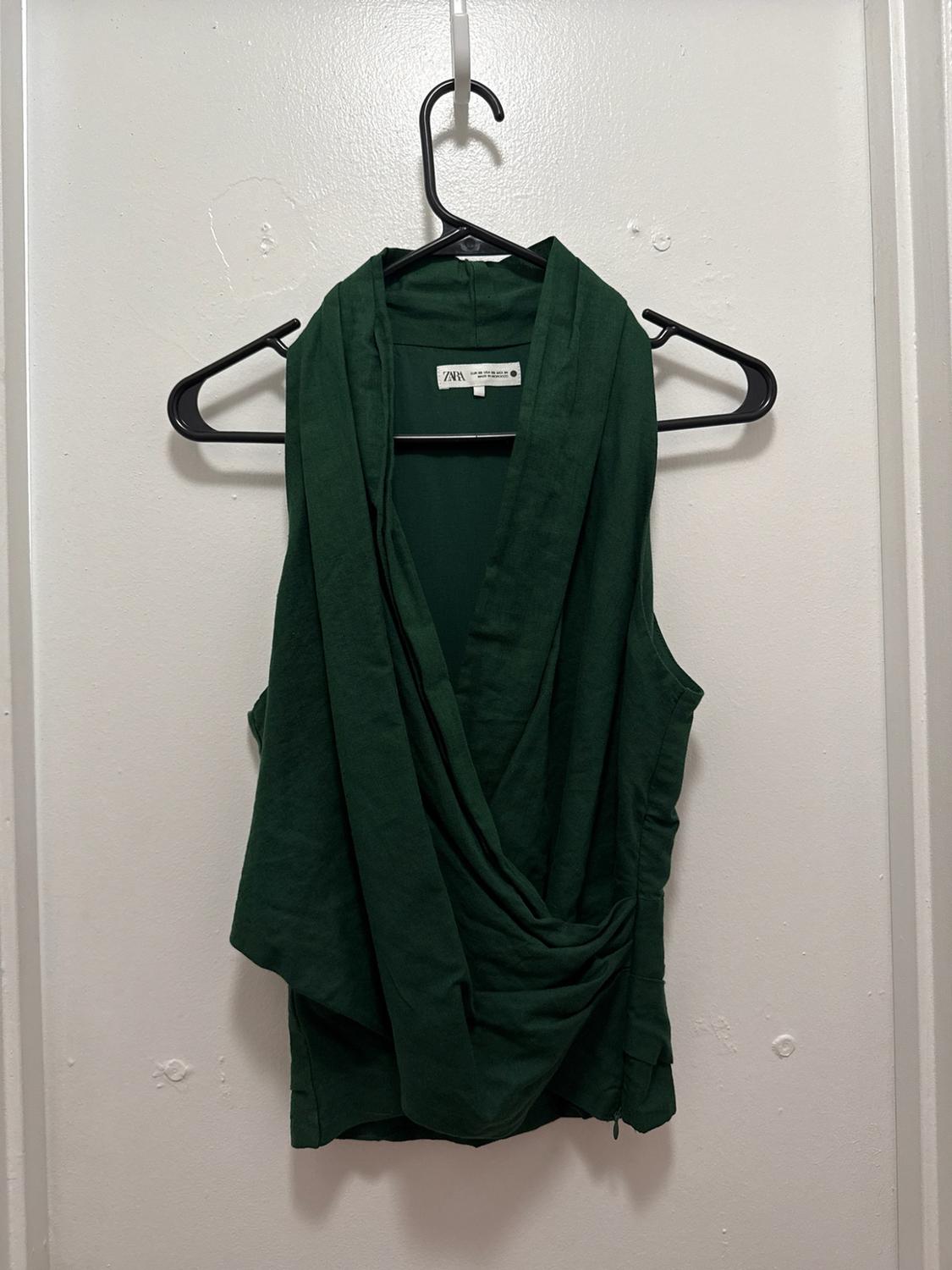 Dark green sleeveless top with a cross-over draped design, hanging on a black hanger against a white background. ZARA text on the label.