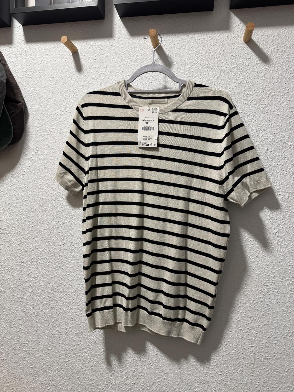 Black and white striped T-shirt hanging on a white wall, with visible tag. Wooden hangers and pictures in the background.
