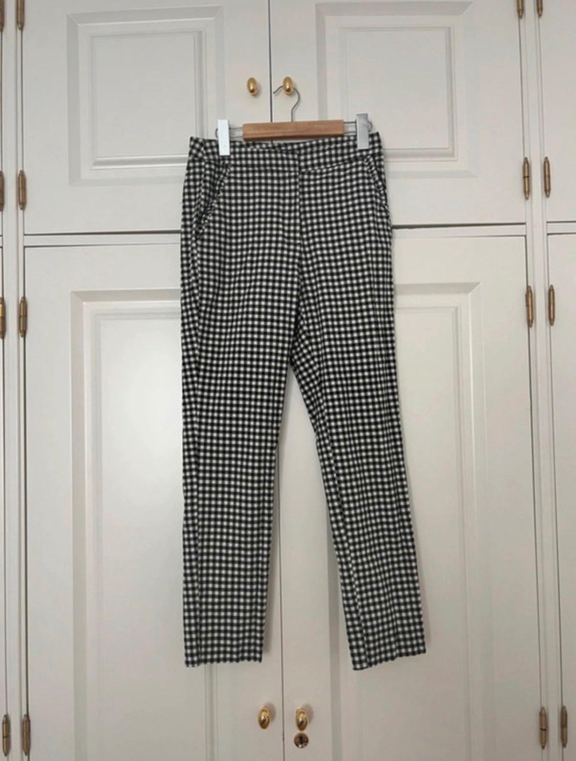 Black and white checked trousers on a wooden hanger, hanging in a white wardrobe with gold handles. Relaxed atmosphere.