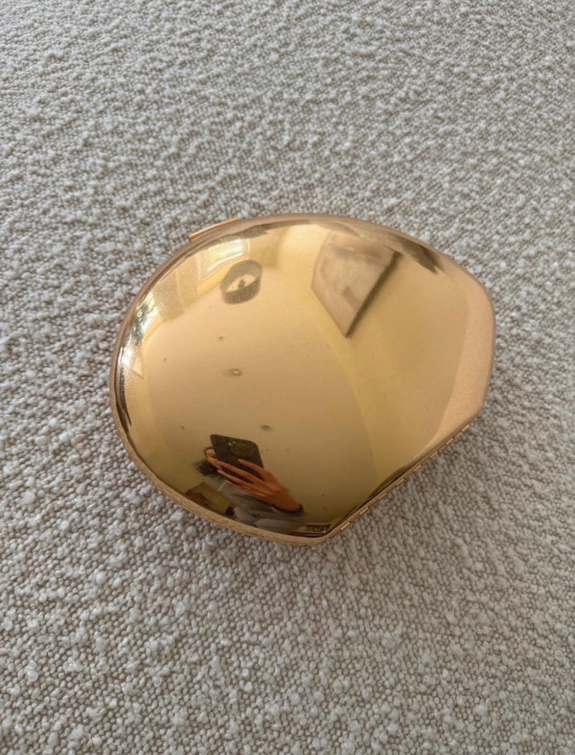 Shiny gold metallic hard clutch bag on a light textured surface. Its reflection shows a person taking the photo with a mobile phone.