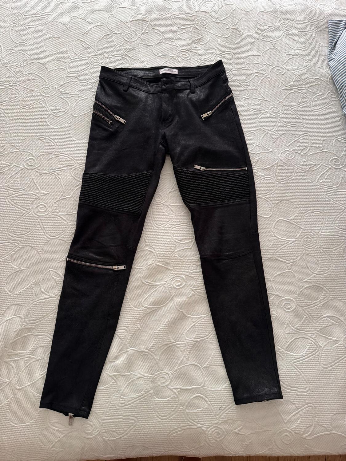 Black biker-style trousers with silver zips and ribbed panels, laid out on a white bedspread with a floral texture.