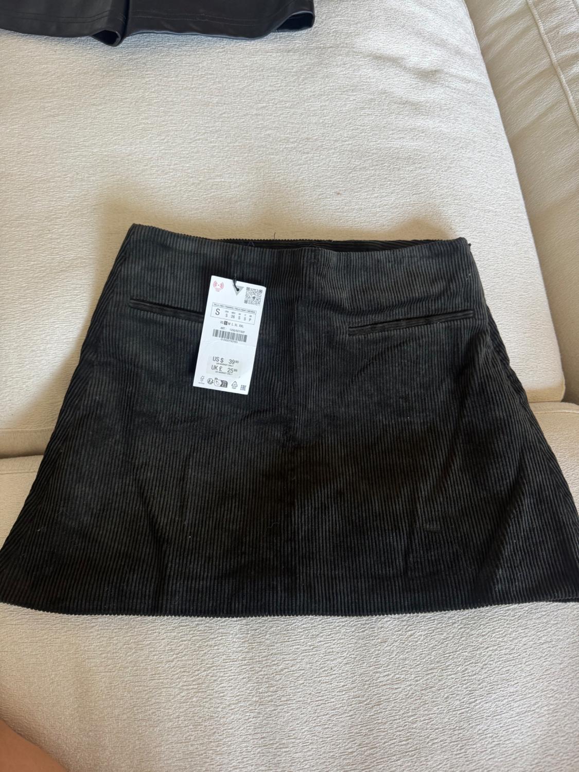 Black corduroy mini skirt with visible tag on a beige sofa. Part of another black garment is visible in the top left corner.