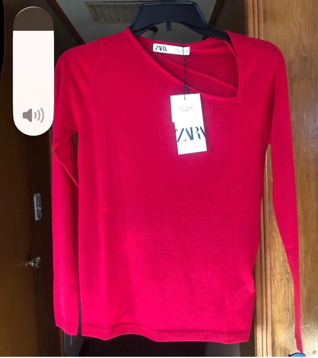 Red long-sleeved sweater hanging on a black hanger, with a visible ZARA brand tag. Wooden door background.