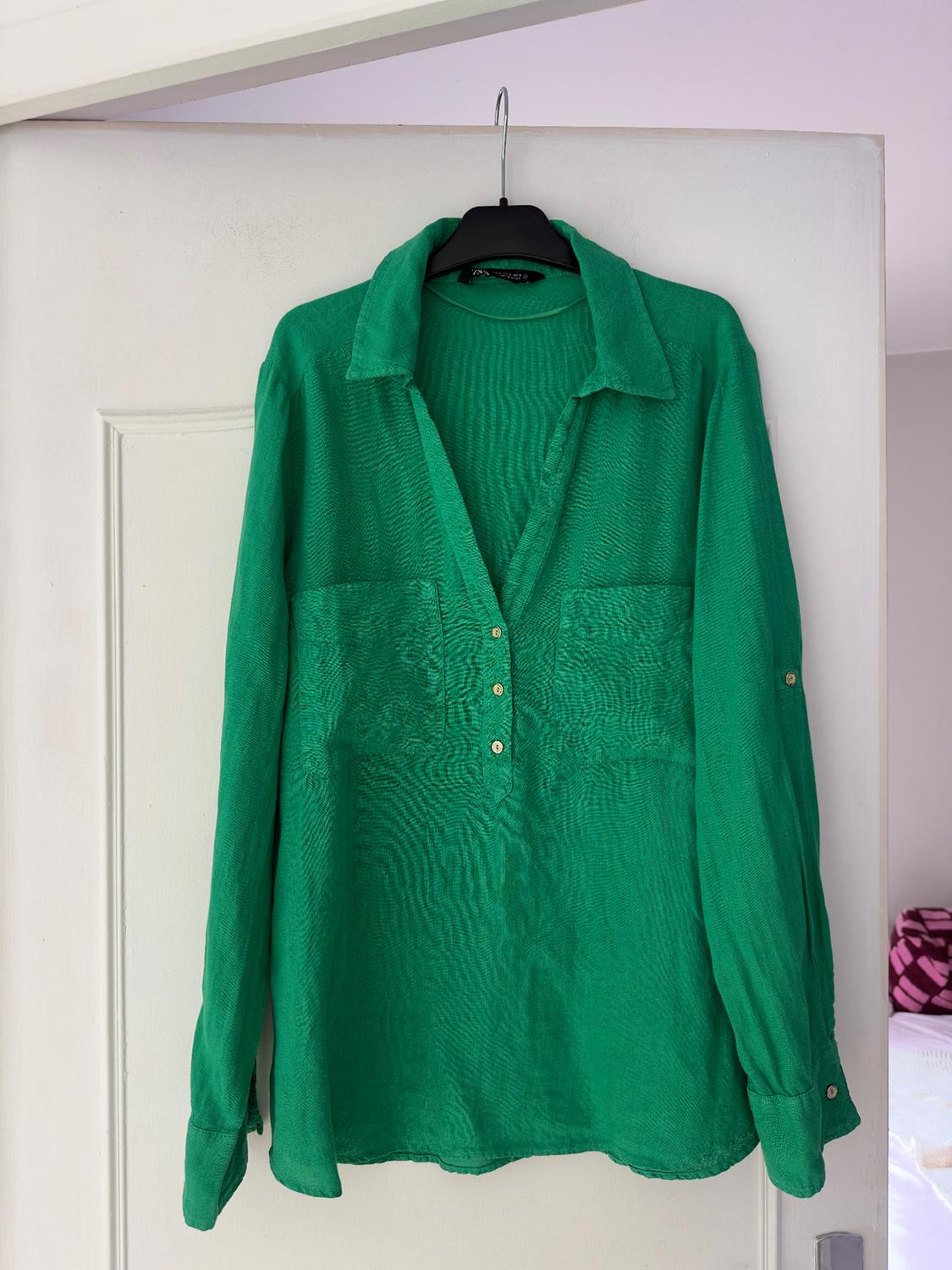 Long-sleeved green shirt hanging on a white door. It has a classic collar and buttons at the top. Indoor setting.