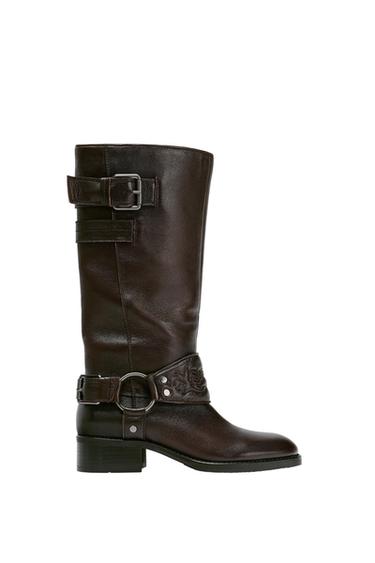 Dark brown leather knee-high boot with a harness detail, metal ring, strap with a floral engraving and buckle at the top.