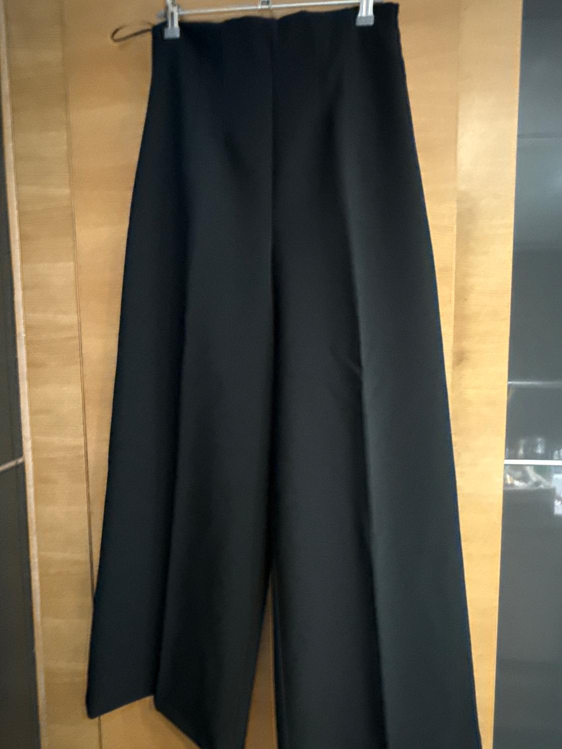 Black wide-leg trousers hanging on a hanger against a light wooden background.