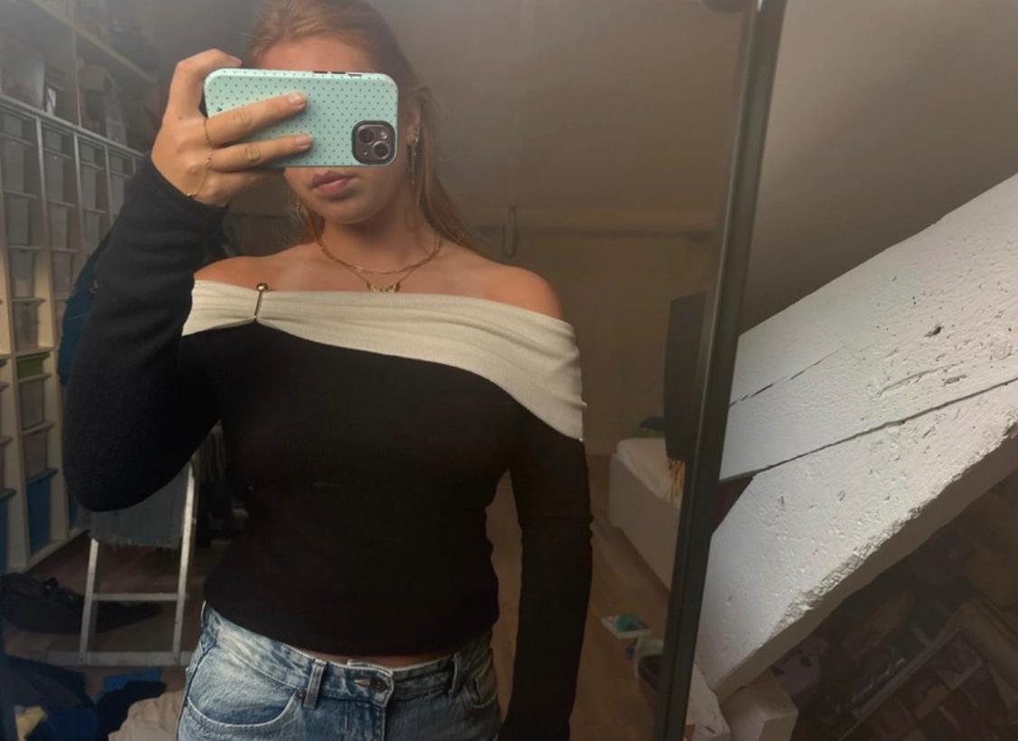 Γυναίκα σε selfie καθρέφτη με μαύρο τοπ, λευκό off-the-shoulder ντεκολτέ, μπλε τζιν και πράσινη θήκη κινητού.