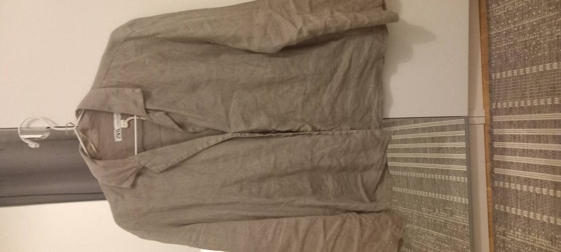 Beige long-sleeved shirt on a white hanger. Inner label with the text ZARA. White door background and striped rug.