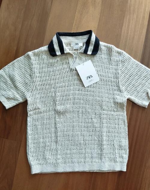 White knitted polo shirt with a blue and white collar, "ZARA" logo swing tag, on a wooden floor.