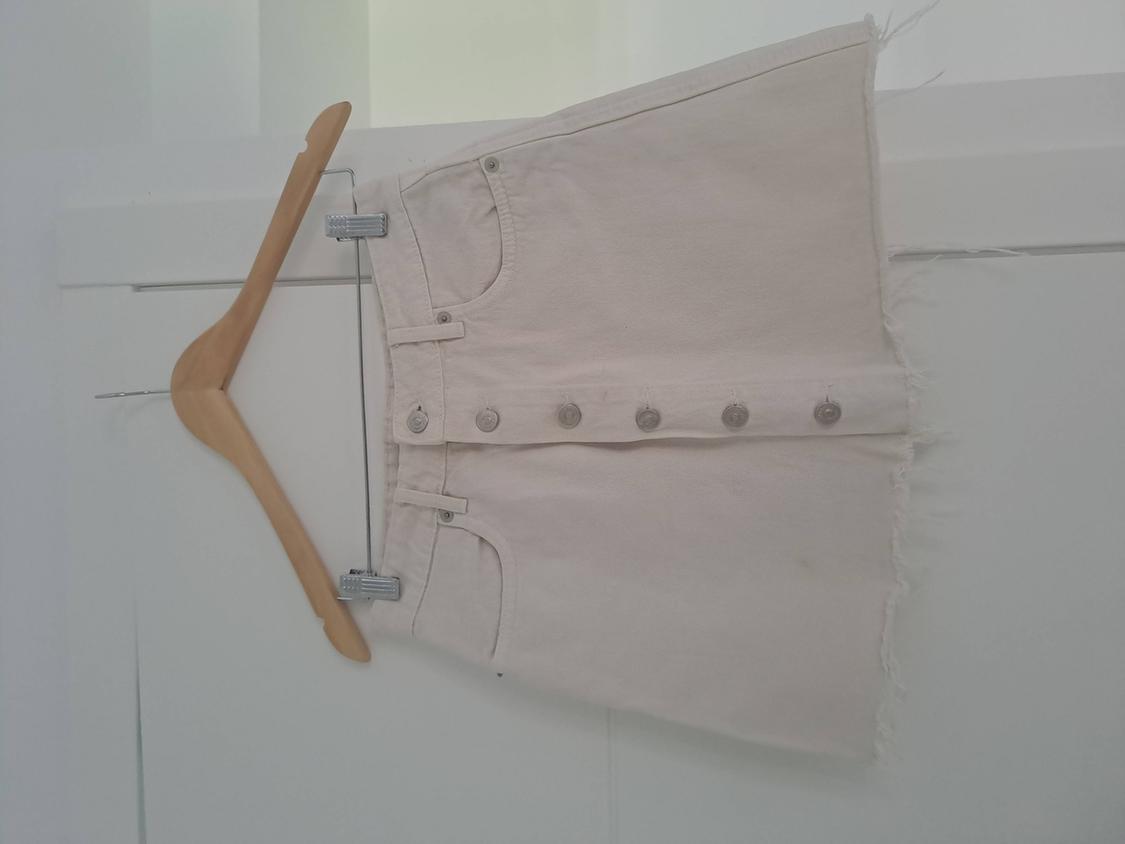Beige denim mini skirt with front metal buttons and frayed hem, hanging on a wooden hanger against a white background.
