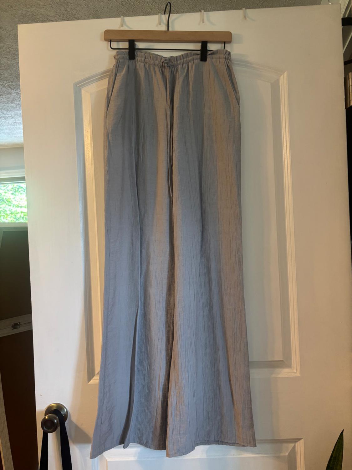 Light grey wide-leg trousers with a crinkled texture, elasticated waist and drawstring, hanging from a wooden hanger on a white door.