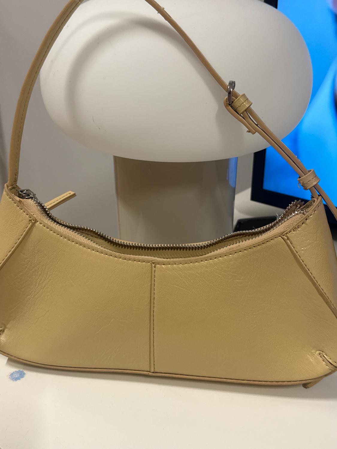 Small beige shoulder bag with zip and visible stitching, on a table next to a white lamp and a blue lampshade.