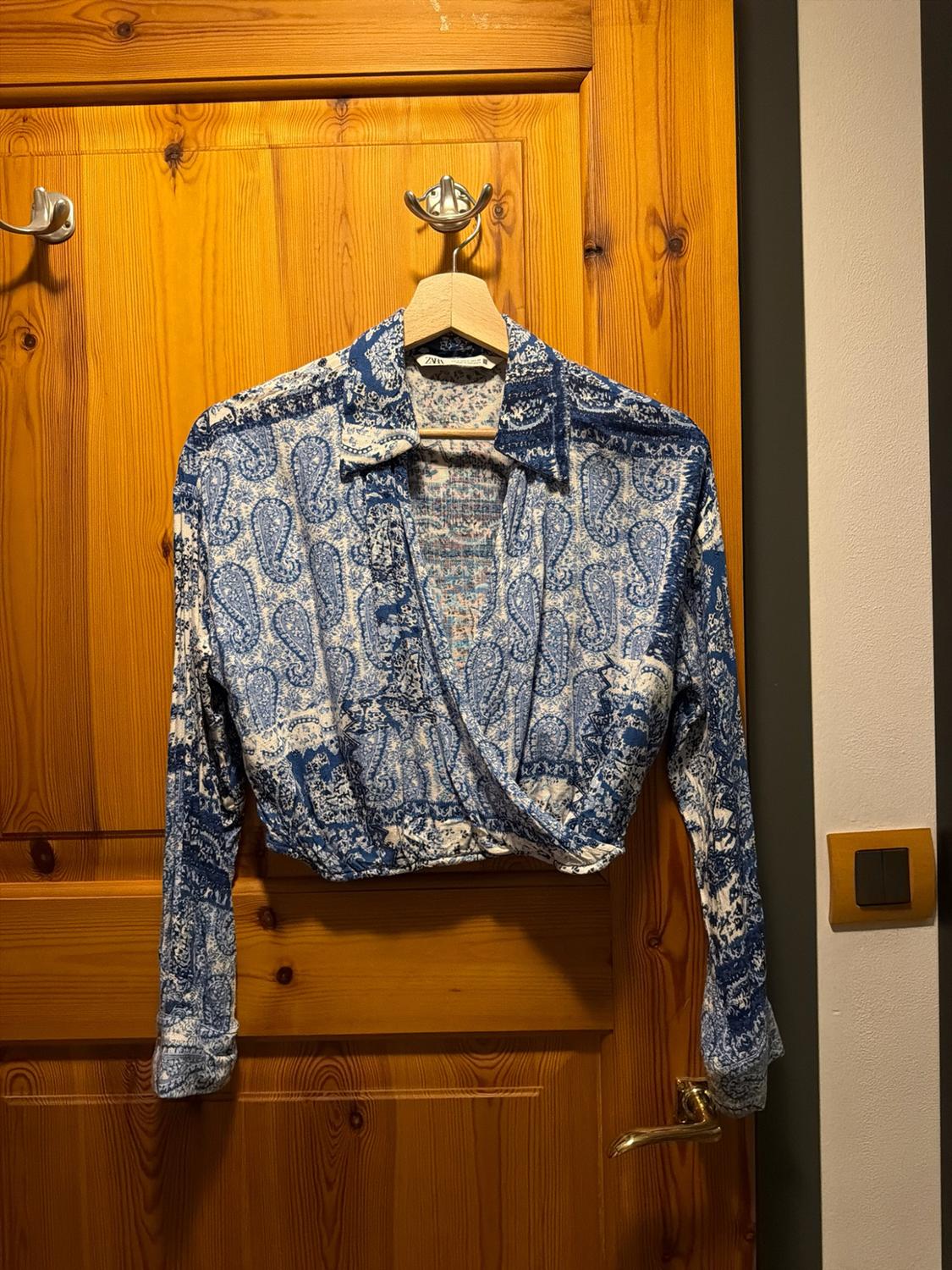 Cropped long-sleeved wrap blouse with a blue and white print, hanging on a wooden door. Label with ZARA text.