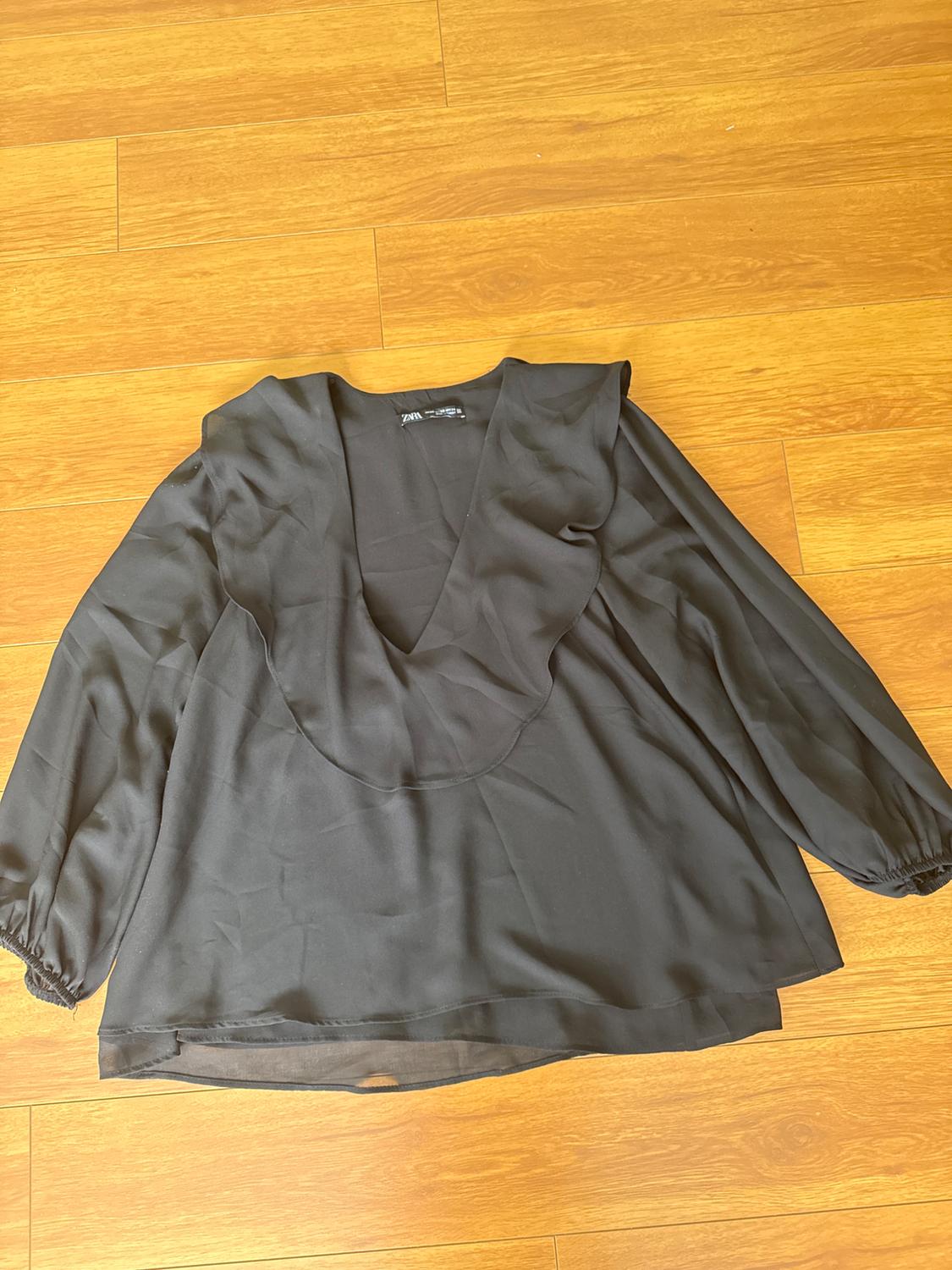 Black long-sleeved V-neck blouse on a light wooden floor. Visible label on the collar.