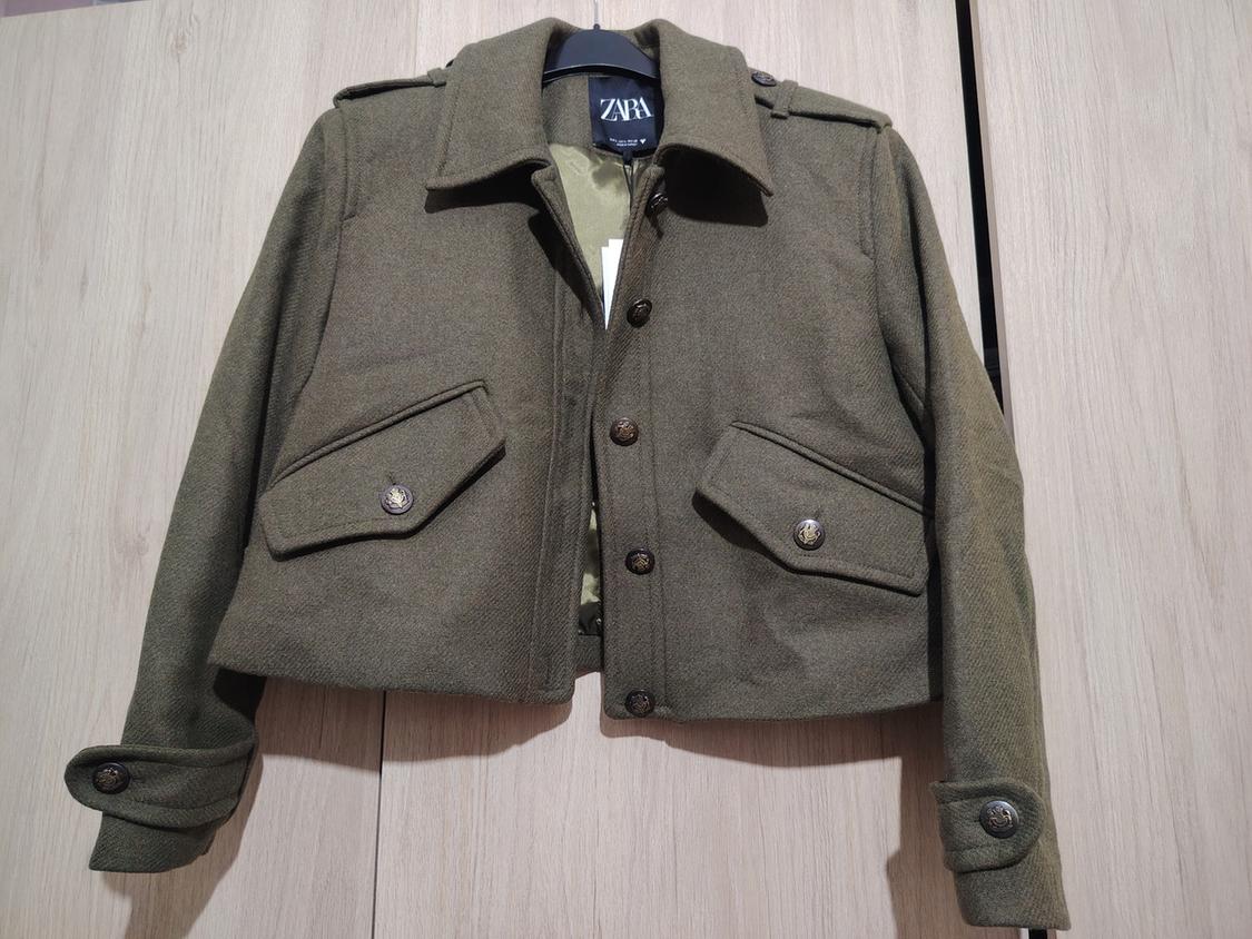 Olive green jacket with buttons and large pockets, hanging on a black hanger with the label visible against a light wood background.