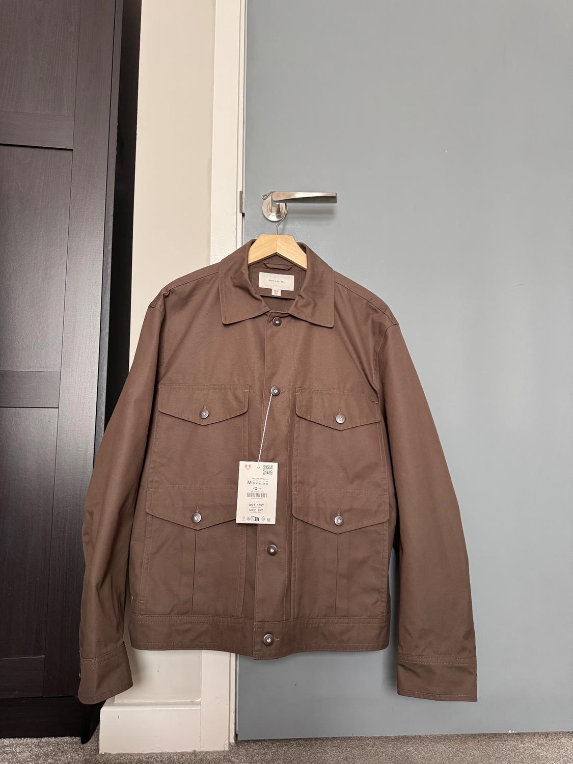 ZARA ORIGINS brown four-pocket jacket, hanging on a wooden hanger on a grey door handle. Label visible.