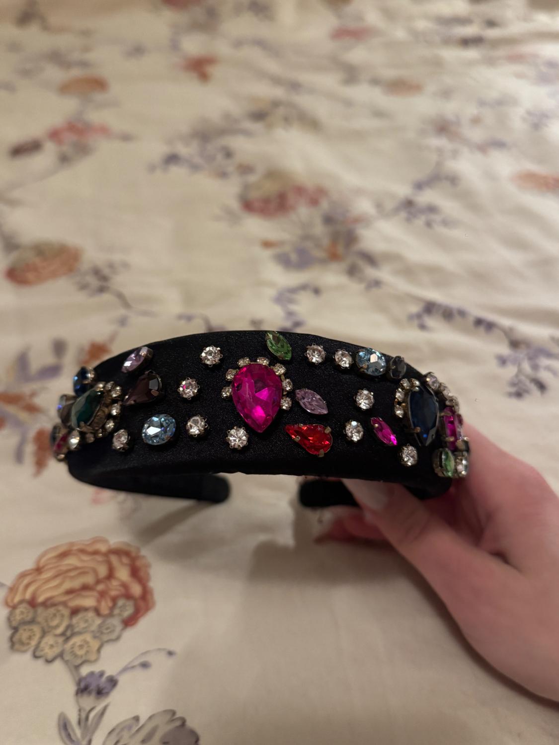 A black headband decorated with vivid colored gems, held by a hand against a floral-patterned fabric background.