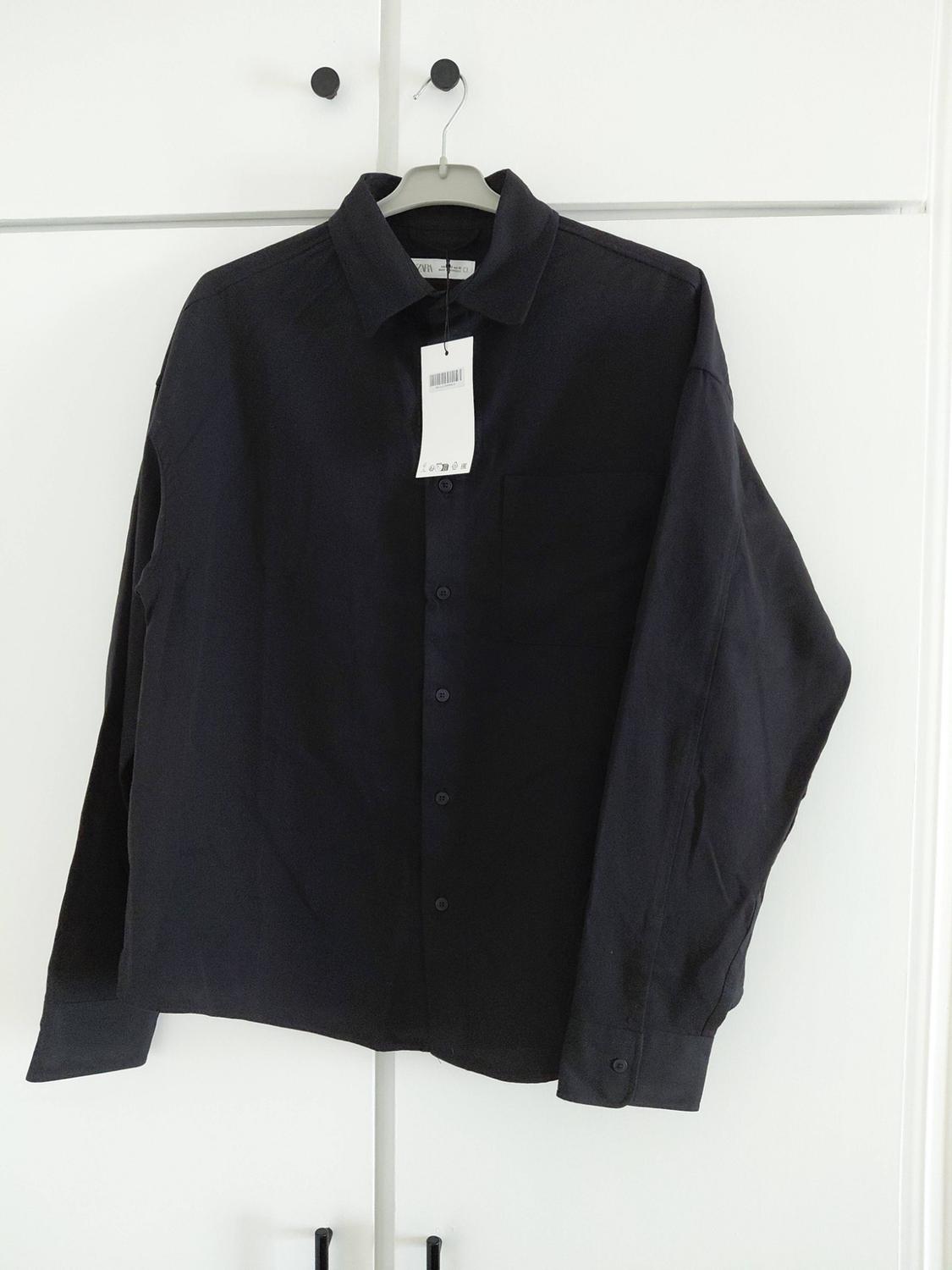 Buttoned black shirt hanging in front of a white wardrobe. It has long sleeves, a pocket, a white label and the text ZARA on the collar.