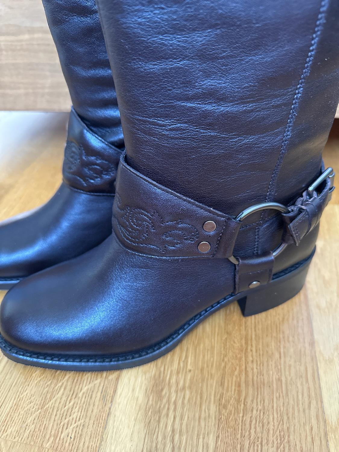Pair of dark brown leather boots with an ankle strap, metal ring and embossed details, resting on a light wood floor.