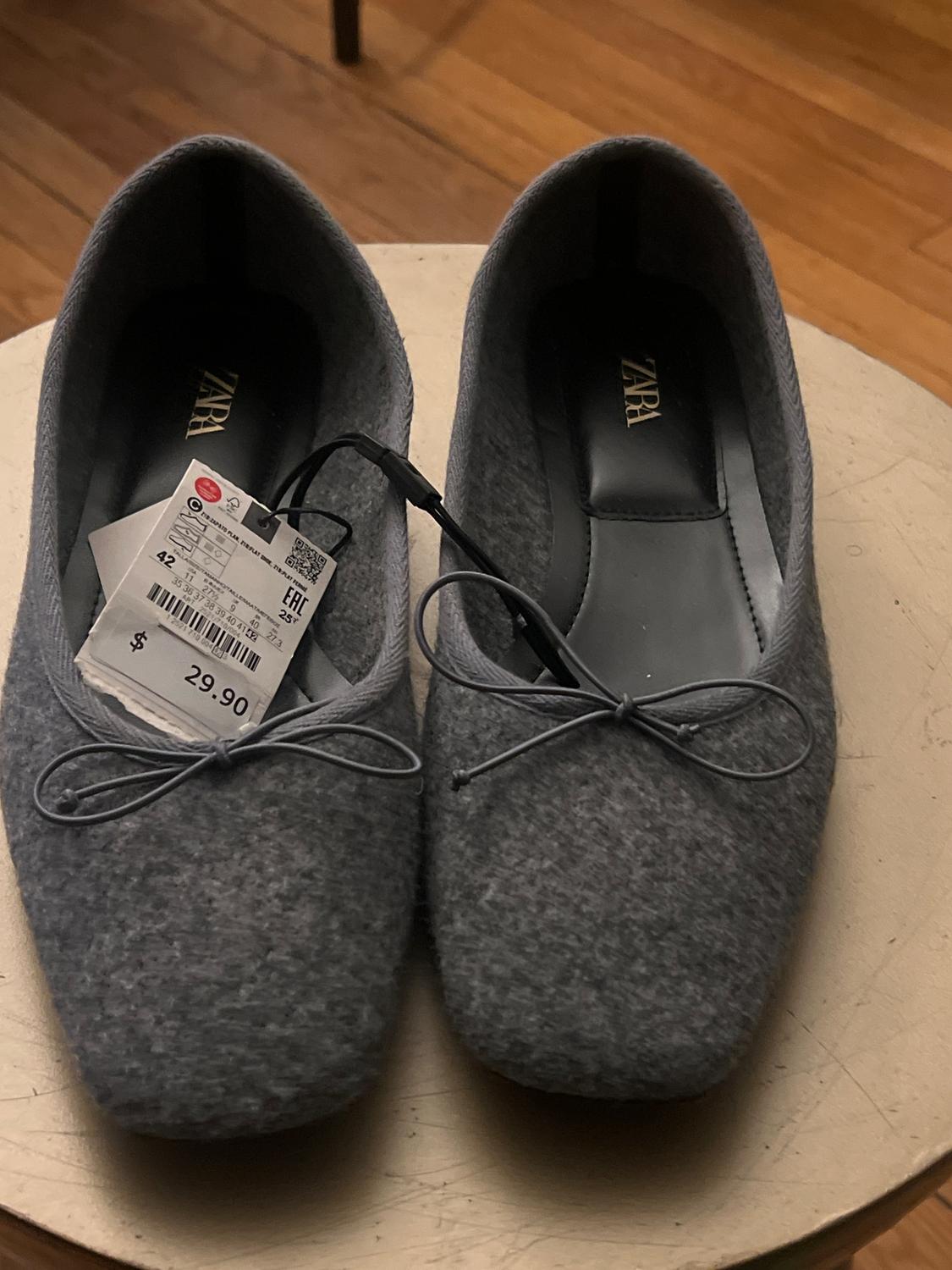 Grey felt shoes with a bow, price tag $29.90, on a round wooden table, wooden floor visible.