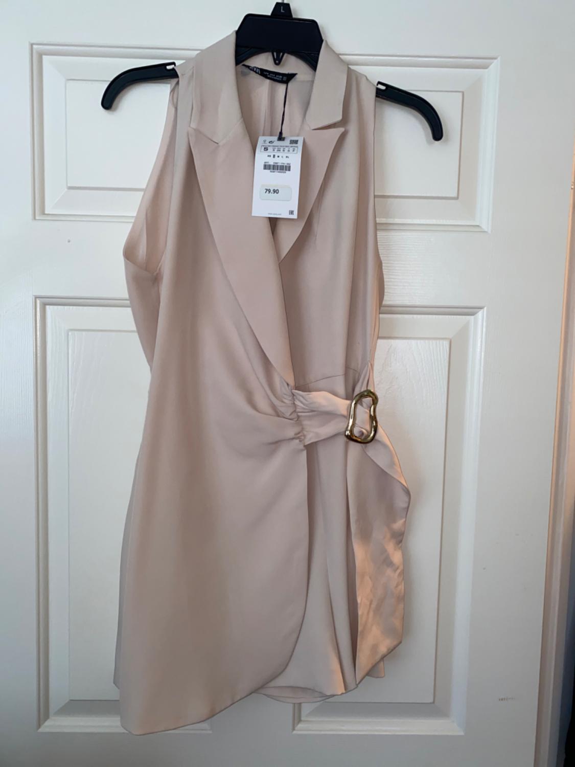 Beige sleeveless dress with gold belt, hanging on a black coat hanger over a white door. Price tag: 79.90.