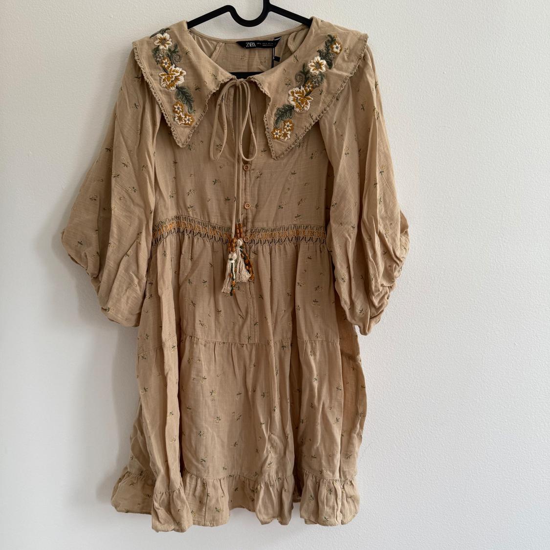 Beige dress with puff sleeves, floral embroidery on the collar, gathered details, and decorative hanging cord at the center.