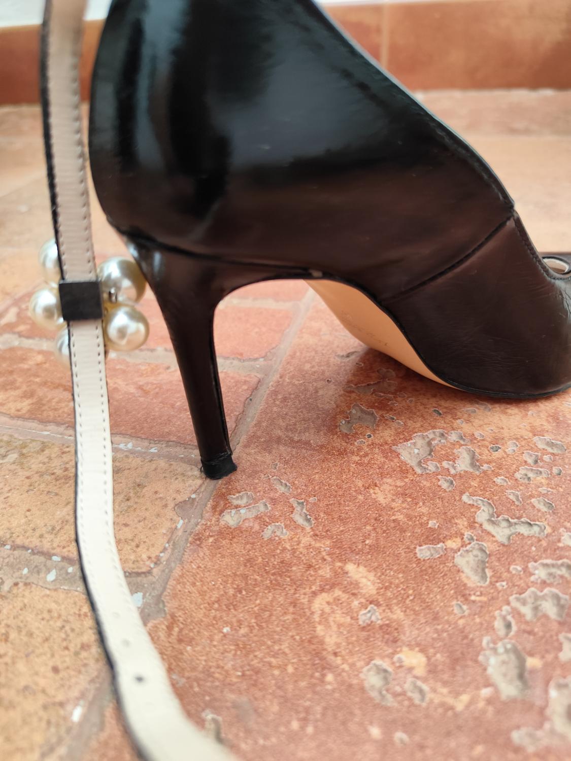 Black high-heeled shoe on a brown tiled floor. Detail of white strap with decorative pearls.