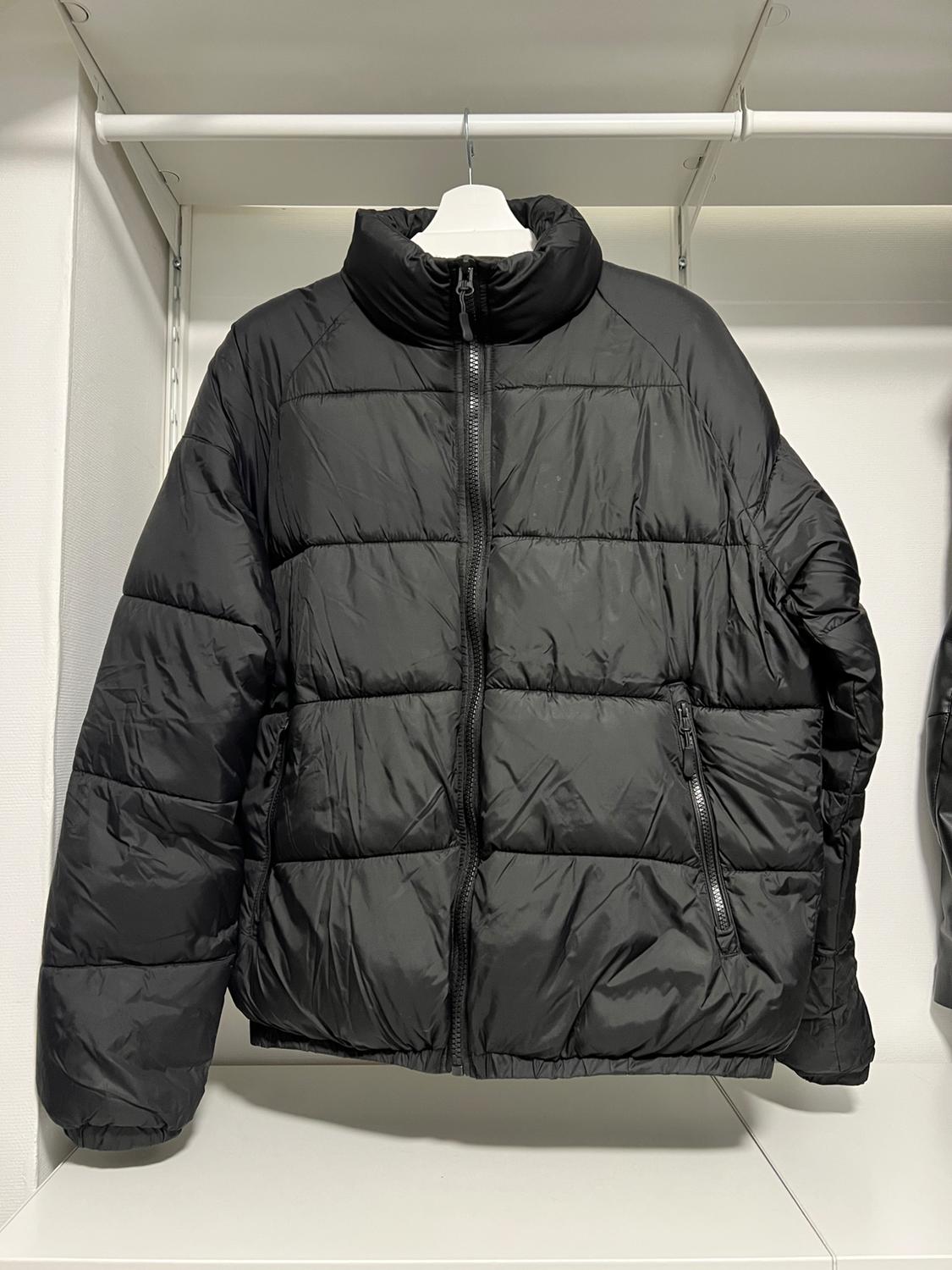 Black padded jacket hanging on a white hanger inside a white wardrobe. It has a front zip and side pockets.