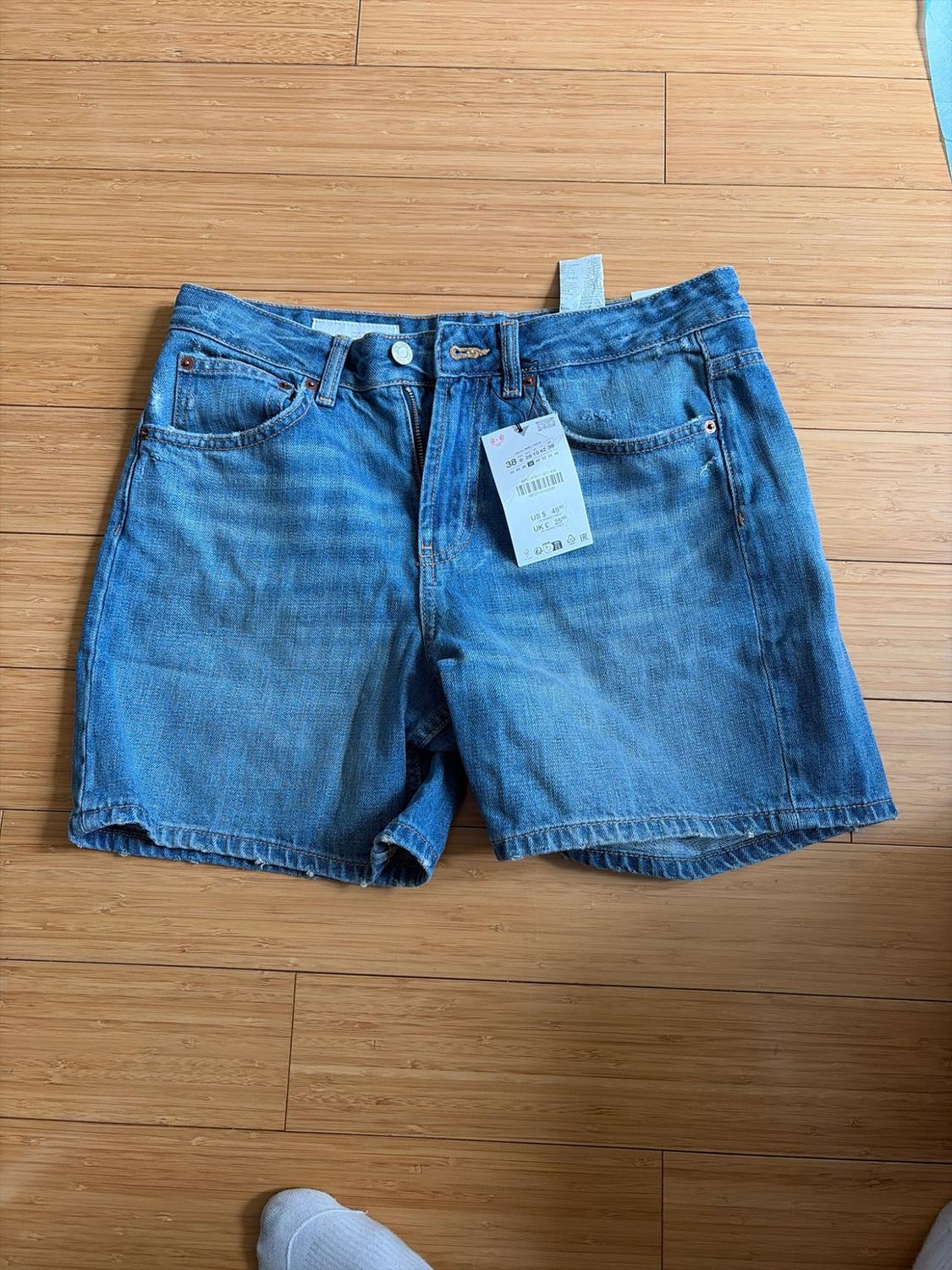 Blue denim shorts with a tag on a wooden floor. A foot with a white sock peeks out at the bottom edge.