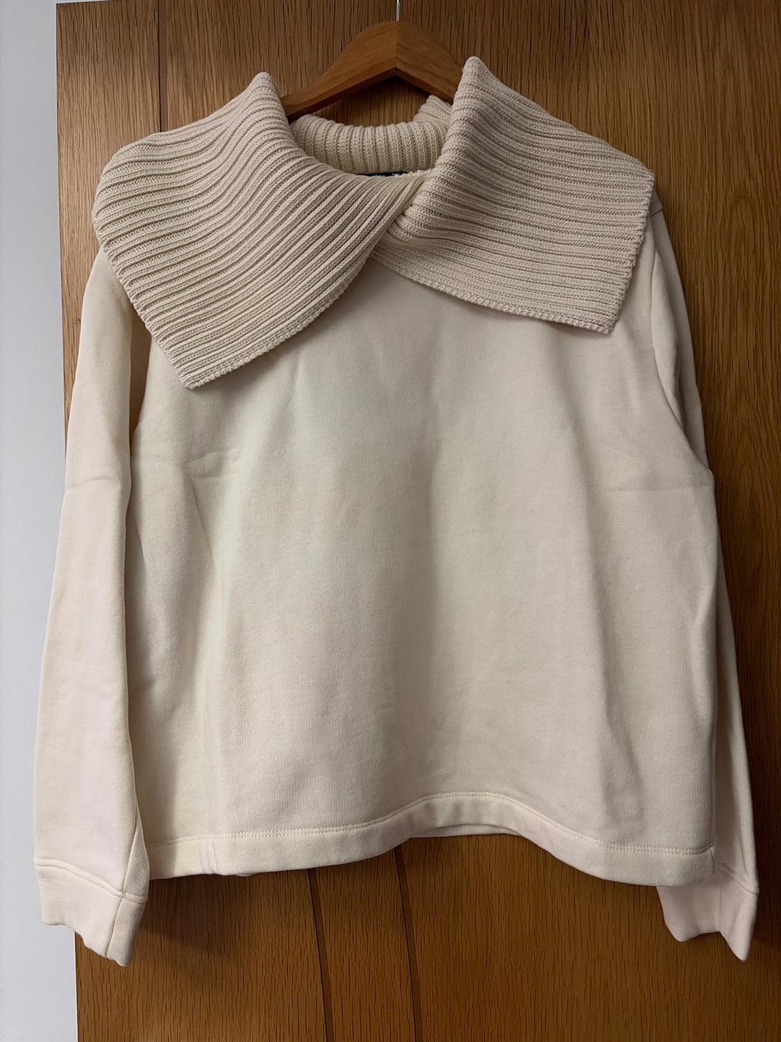 Beige jumper with a wide ribbed knit collar, hanging on a wooden hanger over a light wood door.