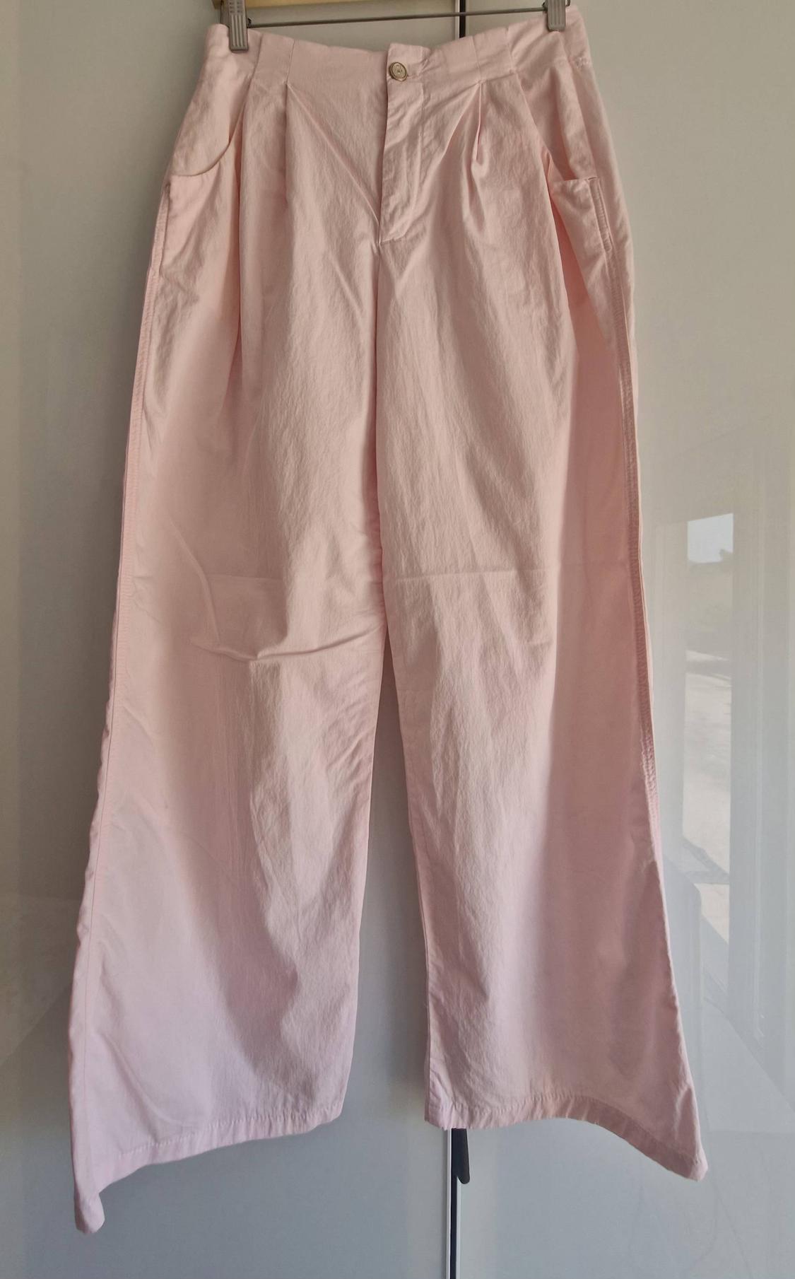 Wide-leg pale pink trousers with pleats and a button fastening, hanging on a hanger against a white background with reflections.