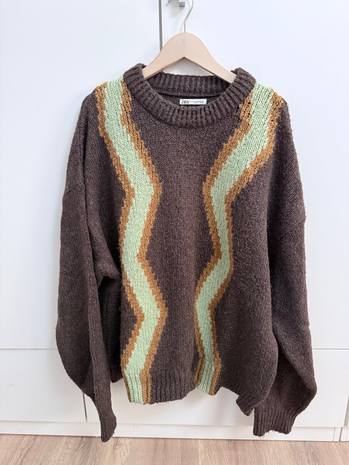 Brown jumper with a green and light brown zigzag design, hanging on a wooden hanger against a white background.